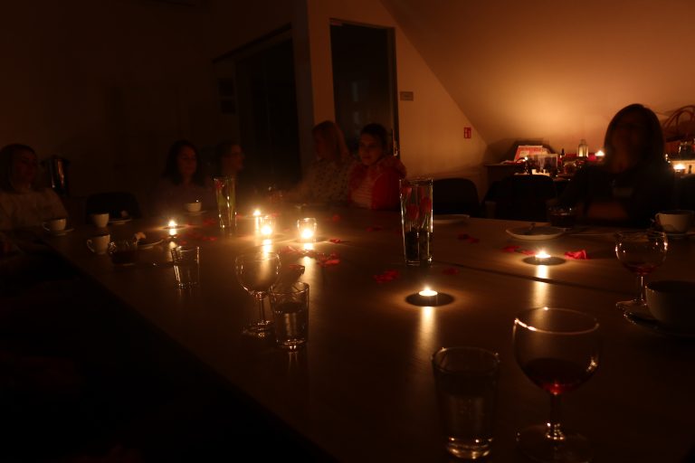 Panie siedzą przy stole, na stole stoją świeczki, jest zgaszone światło.